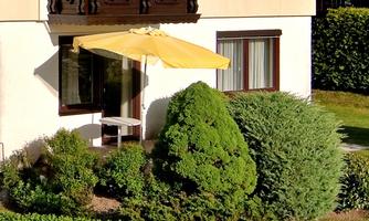 A small garden with green shrubs and a yellow sun umbrella. The windows suggest a cozy house.