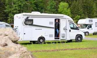 A motorhome is parked at a campsite surrounded by trees. A person is just stepping out of the door of the vehicle.