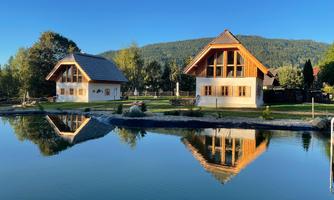 Two charming wooden houses reflect in a calm pond. In the background, green hills rise under a clear blue sky.