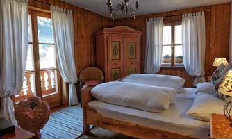 A cozy bedroom with wooden paneling and large windows. The bed is covered with white pillows and blankets, and there is a beautiful wardrobe as well as a lovely seating area.