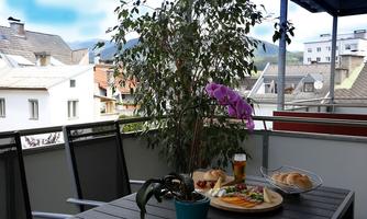 A balcony with a beautiful view of rooftops and mountains. A table is decorated with an assortment of food, a drink, and a blooming orchid.