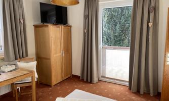 A cozy room with a wooden wardrobe and a table. The large windows let in plenty of daylight, and the curtains are in neutral colors.