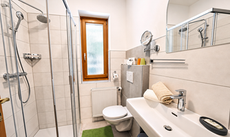 A modern bathroom with a shower and sink. The room is bright and clean, with a small window and a green doormat.