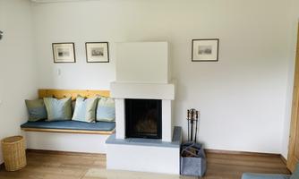 A bright room with a modern fireplace and a cozy bench. Some pictures hang on the wall and a blue armchair stands in the corner.