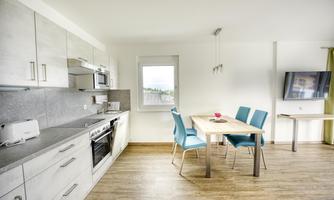 A modern kitchen with bright cabinets and a dining area. A table with blue chairs and a television are also visible.
