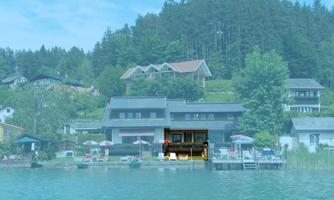 A picturesque shoreline with several houses and green trees in the background. There are parasols and sun loungers by the water, ideal for a relaxing day by the lake.