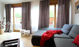 A cozy living room with a gray sofa and a red throw. Large windows with curtains let in plenty of light.
