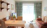 A cozy living room with a dining table, a sofa, and a television.  
Large windows with curtains let in plenty of light.