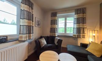 A cozy living room with a black armchair and a gray sofa. The windows let in a lot of light and the curtains have a modern design.