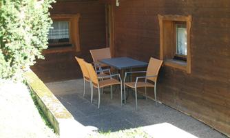 A cozy seating area with a table and chairs in front of a wooden house. The surroundings are green and friendly.