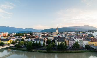 A picturesque view of a town with a river in the foreground and mountains in the background. The buildings are colorful and the church steeple rises into the sky.