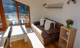 A cozy living room with a brown sofa and many cushions. Large windows let in plenty of light and offer a beautiful view.