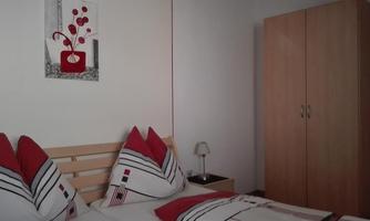 A cozy bedroom with a double bed and red pillows. A decorative picture hangs on the wall, and a wardrobe stands next to it.