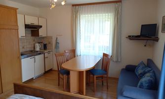 A bright and modern living room with a kitchenette, a dining table, and a cozy sofa. Large windows let in plenty of light.