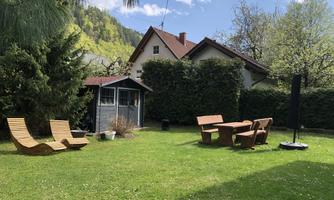A beautiful garden with lawn, wooden lounge chairs, and a table with chairs. In the background, there is a cozy garden house and green trees surround the scene.