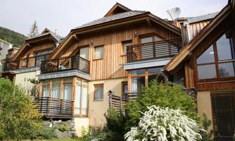 A modern house with a wooden facade and balconies, surrounded by green grass and plants. In the background, trees and hills are visible.