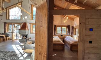 A cozy room with wooden furniture and large windows. In the background, two beds and a fireplace can be seen.