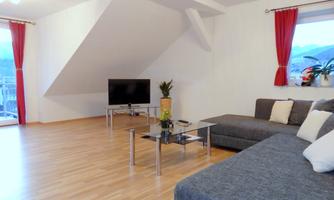 A modern living room with a gray sofa and a television. Large windows let in plenty of light, and there are decorative plants.
