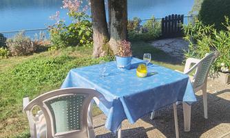 A table with a blue tablecloth is standing outside, surrounded by a garden with a view of a lake. Two chairs and a blue flower pot complete the inviting atmosphere.