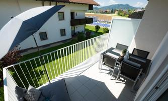 A modern balcony with a dining area and a sun umbrella. In the background, green meadows and mountains are visible.