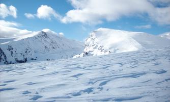 A snow-covered landscape with tall mountains in the background. The sky is blue with some clouds.