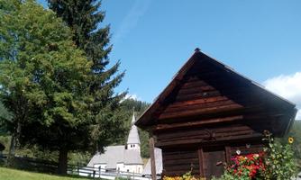 A traditional wooden house stands in a green landscape. In the background, you can see a church tower and blooming flowers.