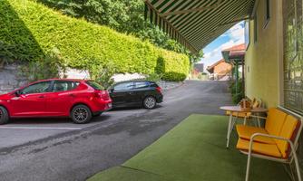 A quiet street with two parked cars, one red and one black. In the foreground is a yellow seating group on a green carpet.