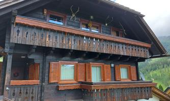 A rustic wooden house with a balcony and windows with wooden shutters. The facade features antlers and colorful floral decorations.
