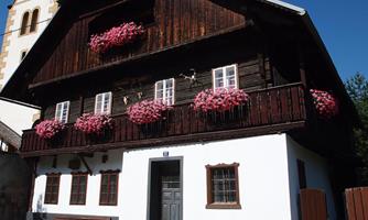 A traditional wooden house with balconies full of colorful flowers. In the background, a church tower is visible.