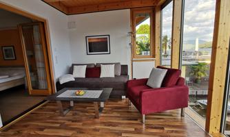 A cozy living room with a brown sofa and a red armchair. Large windows provide a view of the surroundings and let in plenty of light.