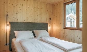 A modern bedroom with two beds and wood paneling. The large window lets in plenty of daylight and offers a view of nature.