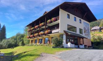 A charming building in alpine style, surrounded by green meadows and trees. Balconies with colorful flowers add an inviting touch to the façade.