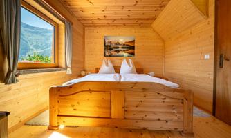 A cozy bedroom with wooden paneling and a double bed. Large windows offer a beautiful view of the mountains.
