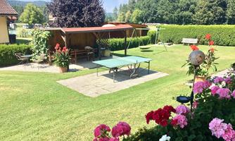 A beautiful garden with a ping pong table and colorful flowers. In the background, there is a wooden gazebo and green grass.