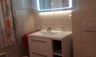 A bathroom with a modern sink and a backlit mirror. There is a shower, a white trash can, and curtains with a floral pattern.