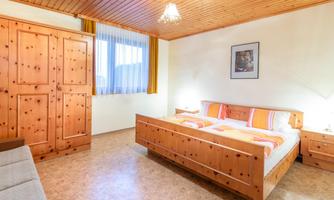 A cozy bedroom with a wooden double bed and orange-white bed covers. The room has a warm, inviting character with wooden paneling and a window that lets in natural light.