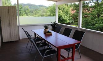 A cozy covered dining area with a long red table and six chairs. In the background, green trees and hills can be seen.
