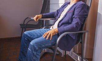A man is sitting relaxed on a balcony in a chair. The sun is shining and casting shadows on the ground.