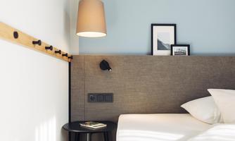 A modern bedroom with a cozy bed and minimalist furnishings. Pictures hang on the wall, and a warmly illuminated lamp creates a pleasant atmosphere.