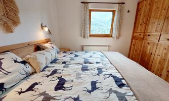 A cozy bedroom with a large bed and a duvet cover featuring a deer pattern. Next to the bed is a wooden wardrobe, and the window offers a view of nature.