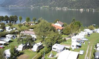 A campsite by the shore of a clear lake, surrounded by trees and mountains. The RVs and tents are arranged on a green lawn.