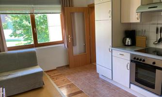 A bright kitchen with a gray sofa and modern kitchen appliances. Large windows provide a view of the outside and plenty of daylight.
