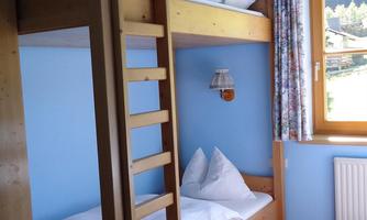 A simple bedroom with a wooden bunk bed and a cozy single bed. The walls are painted light blue and the room has a window with a view.