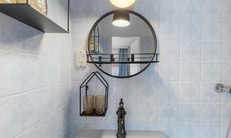 A modern bathroom with a round mirror and an elegant faucet. The walls are decorated with light blue tiles, and in the background, decorative glasses are visible.