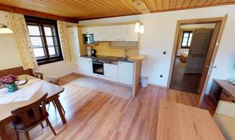 A modern kitchen with wooden flooring and a dining table. In the background, an adjoining bedroom is visible.