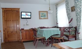 A cozy dining area with a table and chairs. A television hangs on the wall, and the room has large windows with curtains.