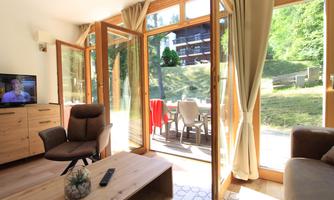 A cozy living space with large windows overlooking nature. The room features a television, an armchair, and a coffee table.