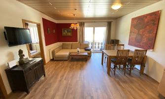 A cozy living room with a sofa, a dining table, and beautiful wooden flooring. The room has large windows that let in plenty of light.