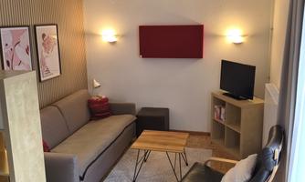 A cozy living room with a gray sofa and a wooden table. Artworks hang on the wall, and a TV is placed on a shelf.