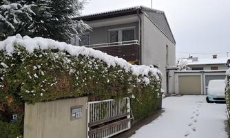 A snow-covered path leads to a modern house. The garden and the hedge are also covered with snow.